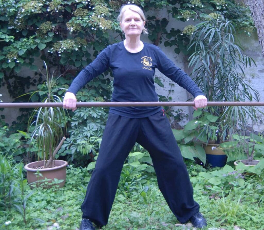 A smiling woman practicing Chinese Yoga with a stick in her garden